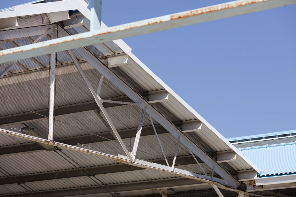 Corrugated metal sheets are supported by thin steel beams, painted white with tinges of rust seeping in against a pale blue sky