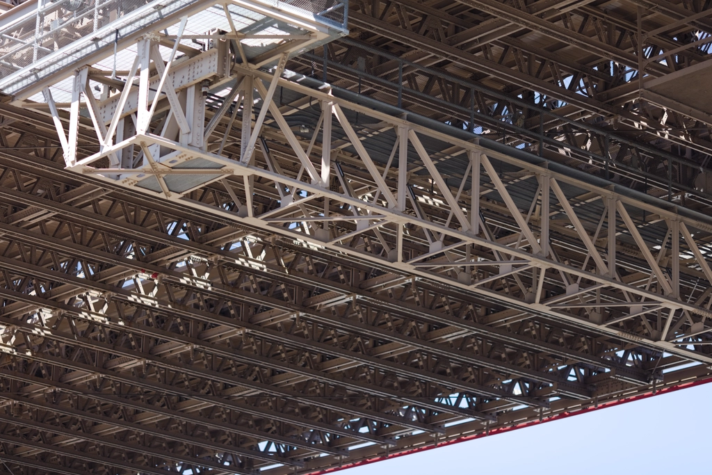 White-and-brown metal latticework forms a sturdy underside to a bridge. The light bounces from somewhere onto it, forming interesting patterns.