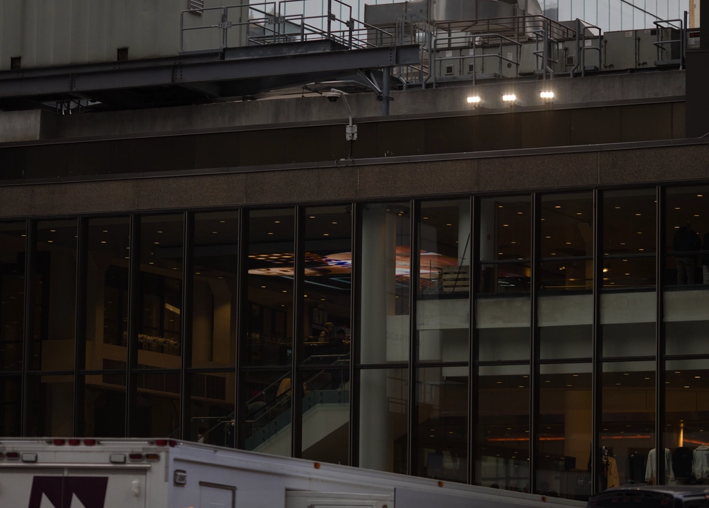 A back door to some building (evidenced by an exposed HVAC unit up top & various pipes) nonetheless has full-story glass windows & nice interior lighting. Various metal railings and concrete pillars both inside and out blend together in the window panes.