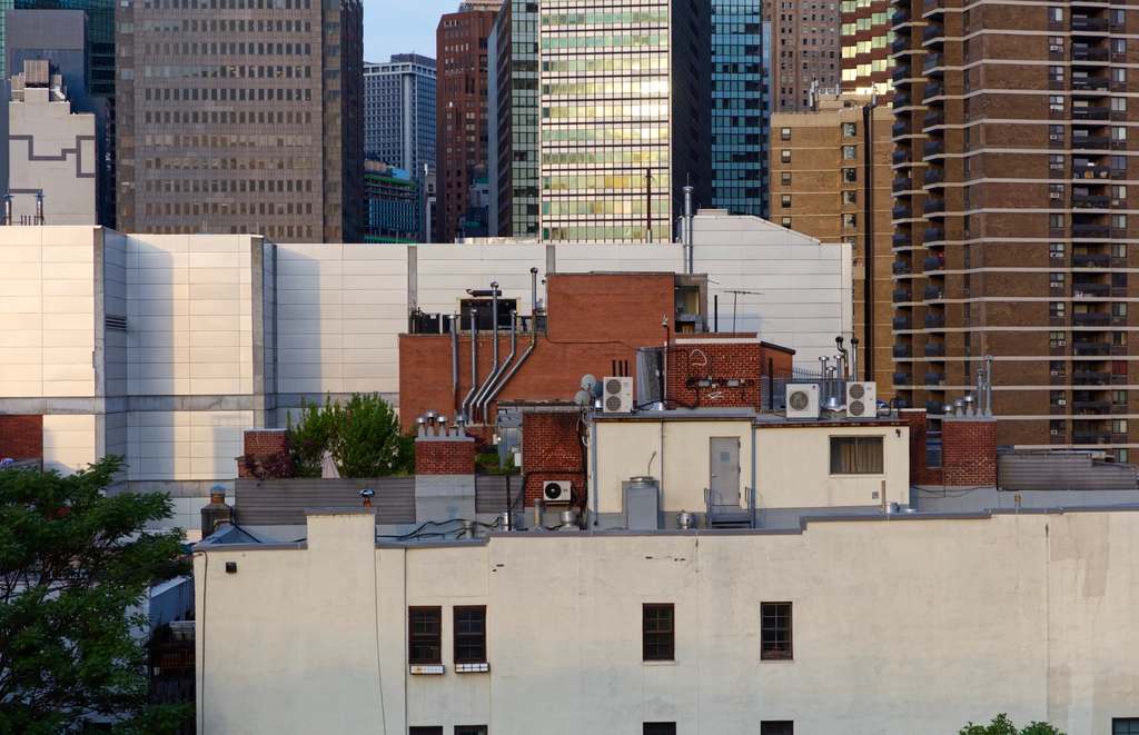A small rooftop area containing a small garden and plenty of AC units, sits tilted in front of a sound wall, in front of myriad skyscrapers, all at slightly different angles. The morning sunlight gives things a peaceful look.
