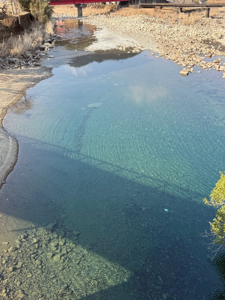 The shadow of a bridge is visible among the rocks & sand at the bottom of a shallow stream, the water colored a beautiful shade of cyan. The wind forms small ripples that only barely disturb the surface, refracting the late afternoon sun in soothing patterns.