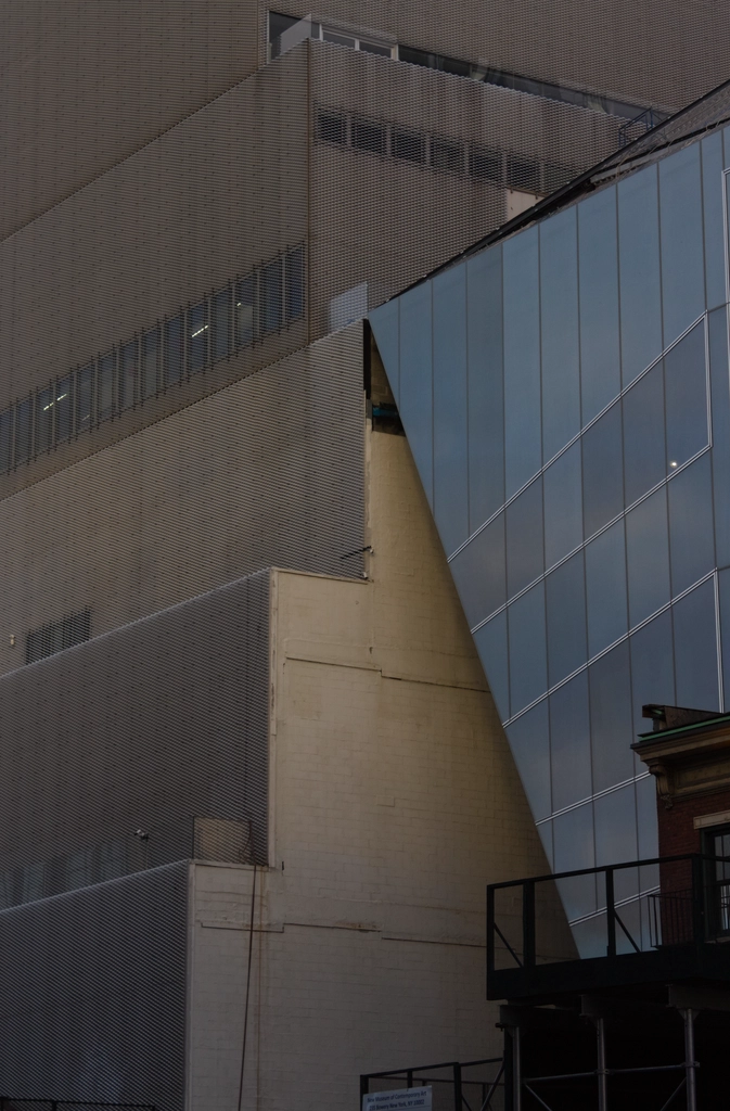 A glass building with fancy angles & custom windows to match runs up right against a fairly older-looking stair-steppy building. The latter is made out of sandy brown painted bricks, with a little bit of facade.