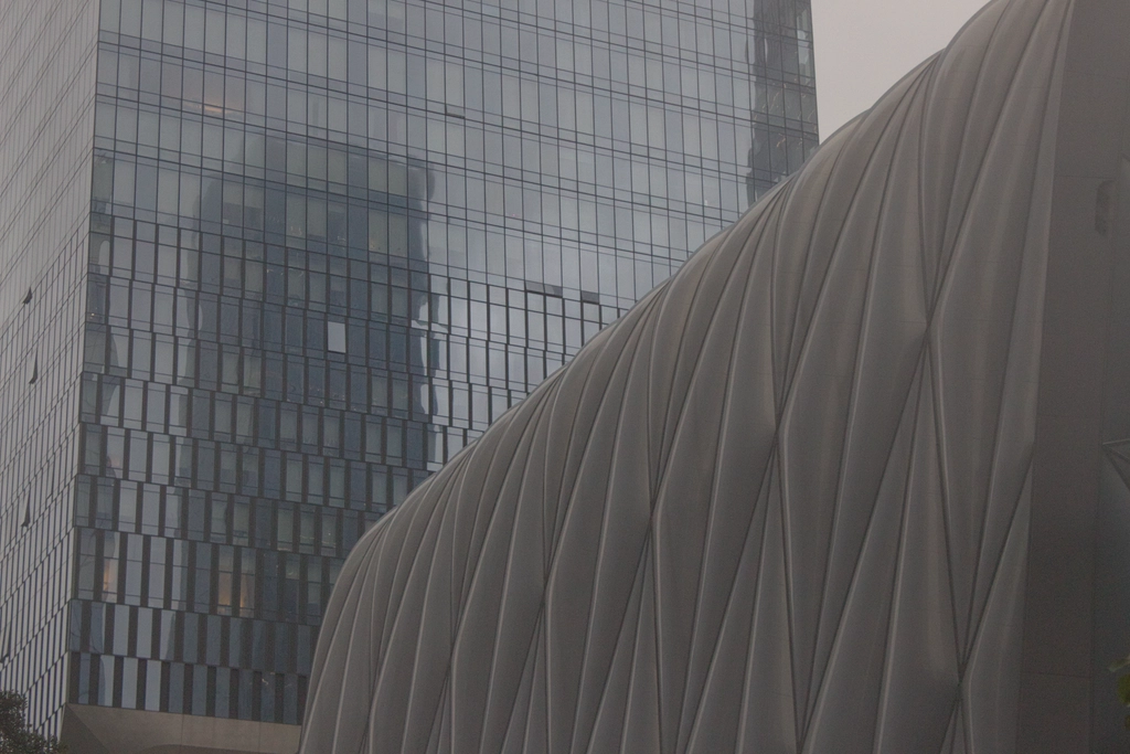 The Shed at Hudson Yards looms in front of some fancy apartment buildings. There is a gray haze over everything making it look dreary.