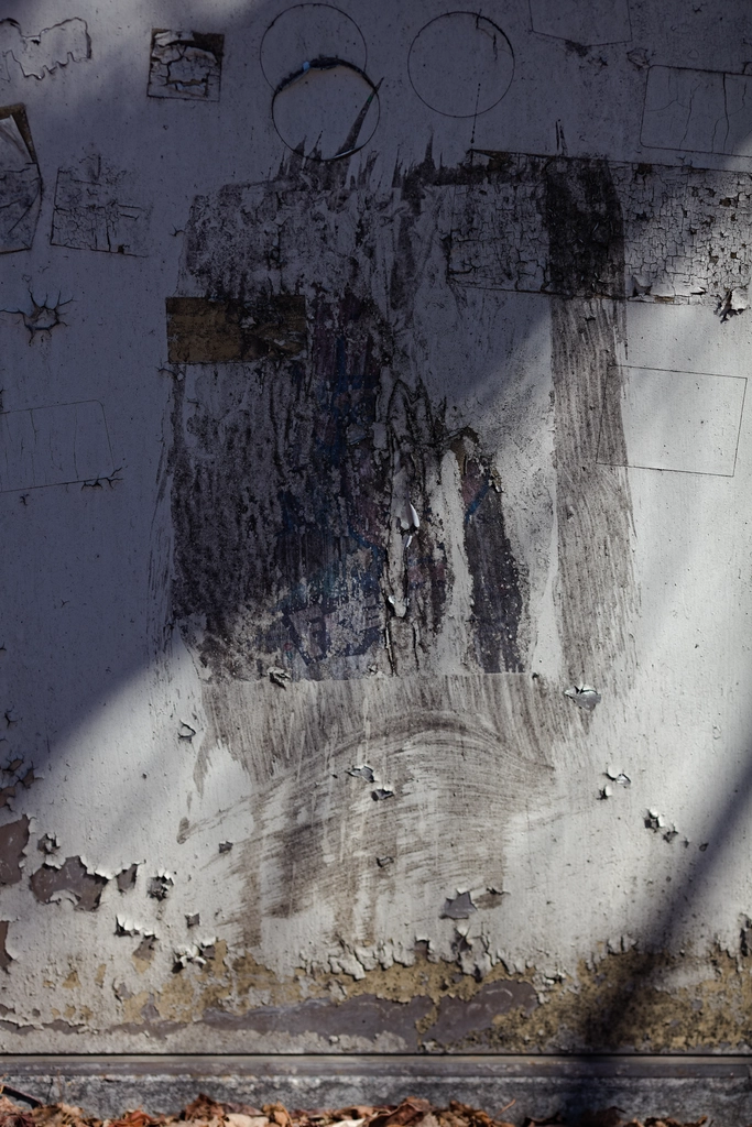 Midday sun casts a diagonal shadow across a light gray wall where a poster once was, before it was sloppily painted over a few times. The rest of the paint is visibly peeling.
