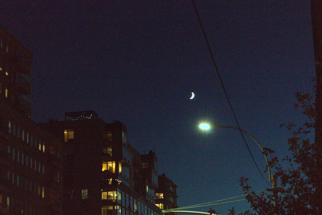 High ISO causes colorful noise to dance across the deep blue night sky, where a small crescent moon hangs above a solitary street light & a few lit apartments.