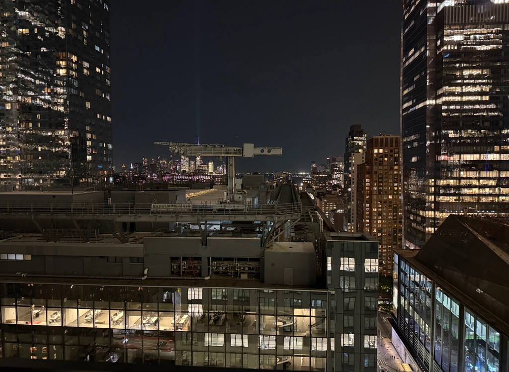 Lots of tall office buildings surround a smaller office buildin's roof, all still lit up despite being fairly late. In the distance, a spotlight pours out from the top of a skyscraper. The city seems to stretch on forever.