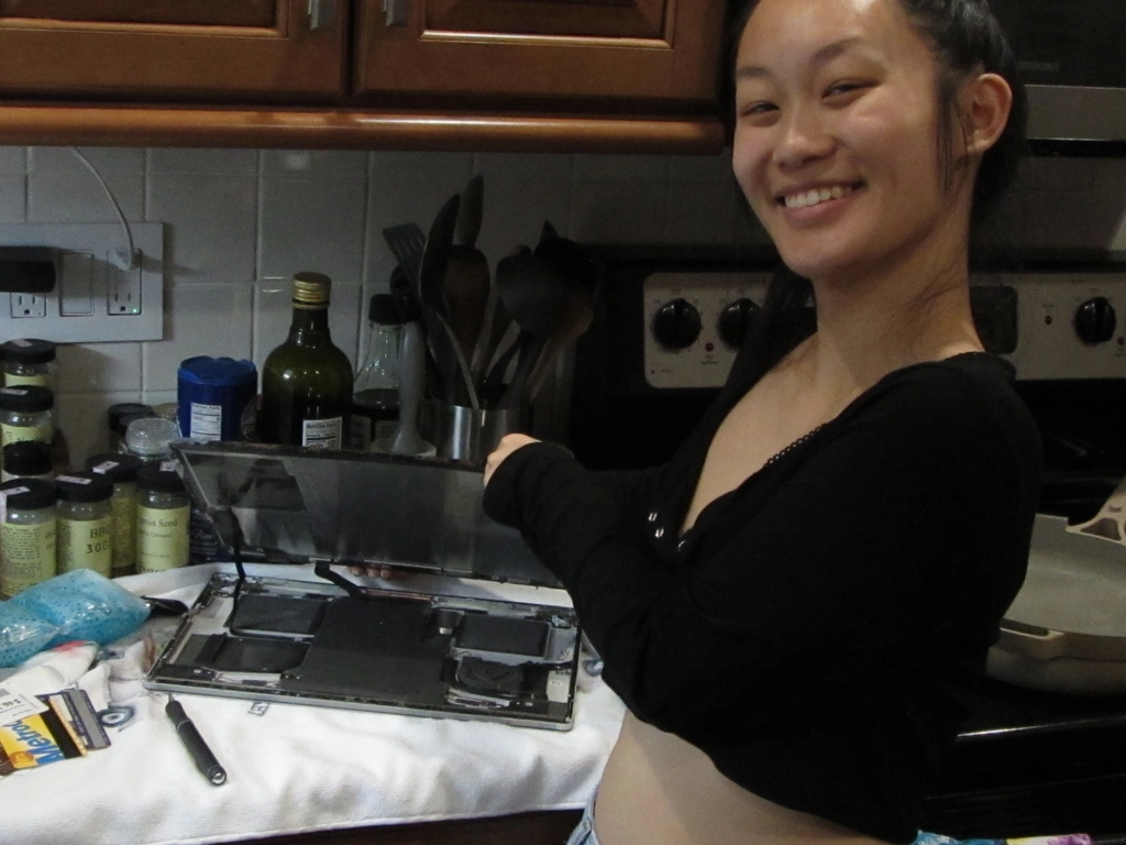 Holly stands holding the laptop screen lifted slightly away from the laptop body, connected only by a few ribbon cables. The cards from the previous step can be seen lying next to the shorn laptop, among spice jars and other kitchen implements.
